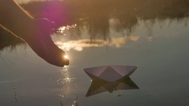 Man with a paper boat in water over beautiful sunset. Man's hand puts paper ship on sea surface. Origami ship Sailing. Dreams, future, freedom or hope concept.