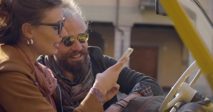 Woman with sunglasses smiling using with smartphone with her boyfriend.Side shot, sun backlight.Caucasian couple in love roadtrip vacation italian travel on convertible vintage car.4k handheld video