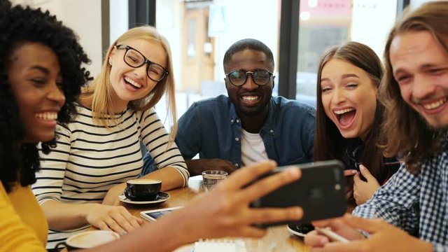 Cool group of young multiethnic friends holding the cell phone to take a selfie in the city coffee shop in the afternoon. Close up shot.