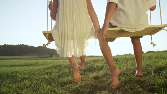 SLOW MOTION CLOSE UP: Unrecognizable kids holding hands and sitting on swing in sunny summer. Barefoot children swinging and watching golden sunset. Young love between boy and girl on romantic evening