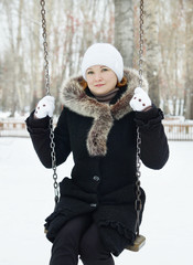 Young pretty woman wearing a black coat sits on the swing in a winter park outdoors