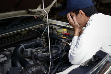 Frustrated stressed young Asian mechanic man in white uniform covering face with hands against car...