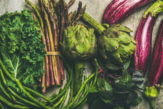 Flat-lay of green and purple vegetables over grey concrete background, top view. Local seasonal produce for healthy cooking. Eggplans, green beans, kale, asparagus, artichoke, basil. Clean eating
