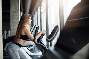 Fototapeta na wymiar Portrait of beautiful female gym attendee running on elliptical cross trainer early in the morning. Workout before everyday job.
