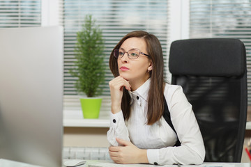 Beautiful serious and engrossed brown-hair business woman in suit and glasses sitting at the desk with tablet, working at computer with modern monitor with documents in light office, looking aside