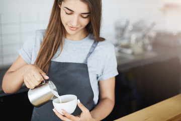 Young professional barista lady pouring milk foam into coffee to create latte art using a jug and a cup. - obrazy, fototapety, plakaty
