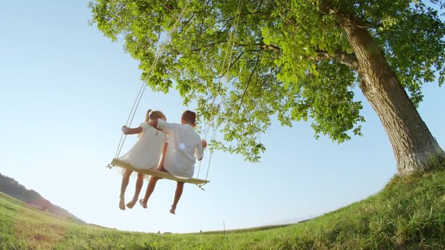 SLOW MOTION Happy boy and girl swaying on swing at golden sunset. Setting sun shining trough couple of smiling kids sitting on big swing. Little brother and sister having fun swinging in sunny evening