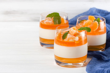 Creamy panna cotta with orange jelly in beautiful glasses, fresh ripe mandarin, blue textile on white wooden background. Delicious Italian dessert. Closeup photography.Selective focus.