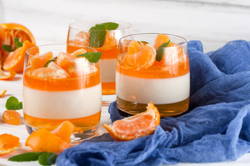 Creamy panna cotta with orange jelly in beautiful glasses, fresh ripe mandarin, blue textile on white wooden background. Delicious Italian dessert. Closeup photography.Selective focus.