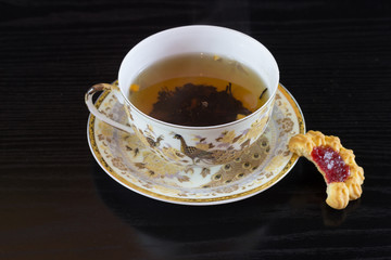  White cup in golden brown patterns of hot tea with homemade biscuit-based saucer  isolated on black background 