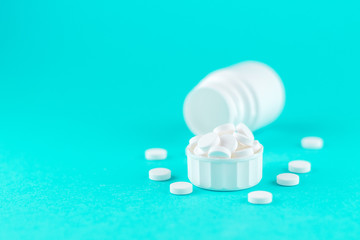Close up white pill bottle with spilled out pills and capsules in cap on turquoise background with copy space. Focus on foreground, soft bokeh. Pharmacy drugstore concept