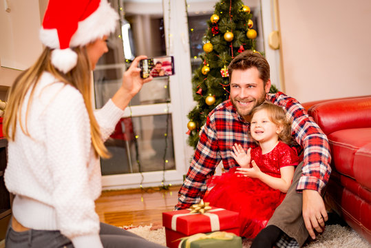 christmas, holidays, family and people concept - mother photographing happy father and little daughter with present by smartphone