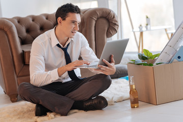 Fototapeta na wymiar Interested man. Calm cheerful young man looking at the screen of his laptop while sitting on the floor with his legs crossed while a bottle of alcohol and a box of personal items standing by his side