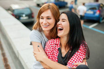 Two young sisters hugging, laughting and sitting near road in the street. one brunette girl in red plaid shirt, another redhead girl wearing gray shirt and blue skirt. concept of sincere friendship