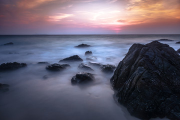 Fototapeta na wymiar Dark rocks in a blue ocean under colorful cloud sky - Long exposure photography