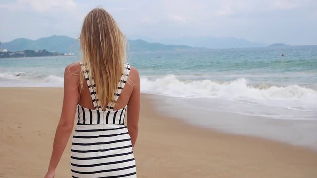 Young smiling woman in black and white dresses walking alone on a tropical beach and looking at camera over background at storm on the sea. Travel Concept.