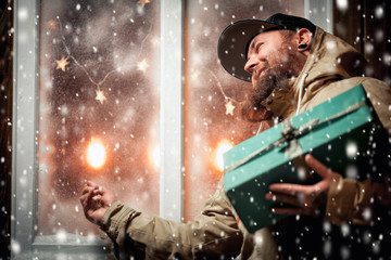 Christmas night, snow falling, funny man with a beard and with gifts in hands by the window. Winter background with lit garlands