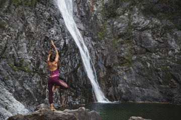 Fototapeta na wymiar Italy, Lecco, woman doing Tree Yoga Pose on a rock near a waterfall