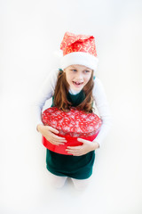 A little girl in a Santa Claus hat on her head and a green sarafan holds a large gift round red box in her hands and smiles.