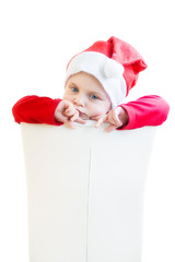 A baby dressed as Santa Claus sits backwards in front of her on a white chair and thinksfully sucks her finger.