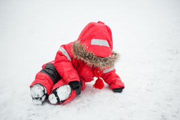 A child in a red jumpsuit sits on white snow and looks down, hiding his face in the hood