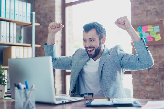 Portrait of young, smiling, attractive finansist, triumphant  having his arms with fists raised, shouting, creaming, celebrating completed work, having his mouth opened with beaming smile