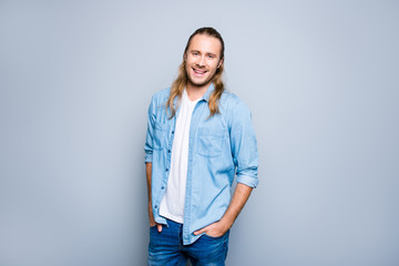 Happy joyful young hipster with long blonde hair is smiling, holding his hands in pockets ans posing against grey background