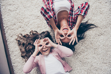 The best time in my life! Top view photo full of fun and laughter. Happy mother wearing checkered shirt and her little daughter are lying on the floor and making binoculars with their fingers