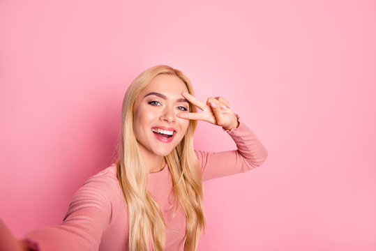 Close up photo of happy adorable woman with beaming smile. She is taking selfie and showing two fingers against pink background