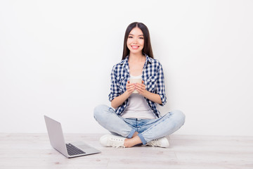 Attractive, cute, charming girl sitting on the floor with crosssed-legs near wall, holding mug of tea in hands, smiling and looking to the camera, having rest from studying on white background