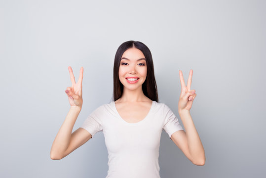 Portrait of glad, funny, smiling, toothy, beautiful girl showing with two hands v-sign, two peace symbols, standing over grey background