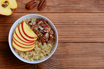 Quinoa porridge with fruits and nuts topping. Vegan quinoa porridge with apples and pecan nuts in a white bowl and on brown wooden background with empty space for text. Healthy breakfast. Top view