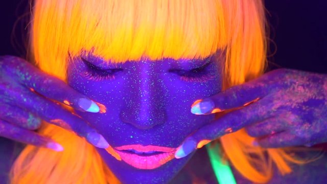 Closeup woman face with fluorescent make up in orange, wig, creative makeup look great for nightclubs. Halloween party, shows and music concept - slow motion video