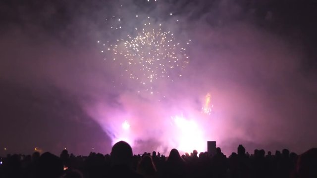 Big crowd of people watching multicolored multicoloured firework display, some taking pictures or filming using their smartphones or mobile phones