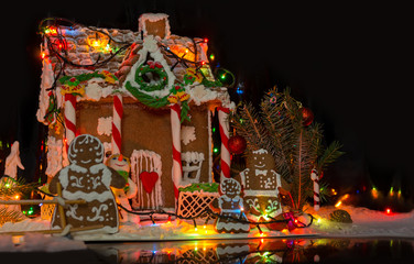 Cute gingerbread family near big snow-covered homemade gingerbread house with Christmas lights on dark background