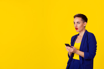 Shocked business woman with phone looking to the side copy space isolated yellow background wall. Business attire formal blue suit, short hair, red lips