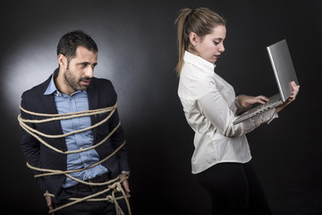un uomo triste in giacca è legato mentre la sua compagna controlla il suo computer - sfondo scuro  - obrazy, fototapety, plakaty
