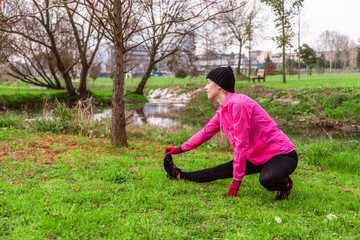 Young woman warming up and stretching the legs before running on a cold winter, autumn of fall day in an urban park. Female athlete wearing pink windbreaker, beanie, gloves and running tights