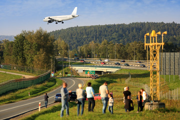Ludzie oglądają lądowanie samolotu pasażerskiego, w tle autostrada.