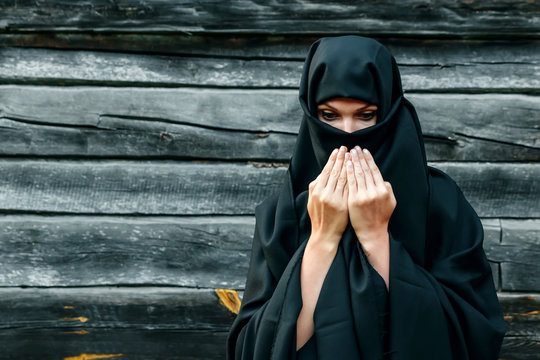 A beautiful, young, Muslim girl in a black veil with a closed face on the background of a gray tree prays. Copy space.