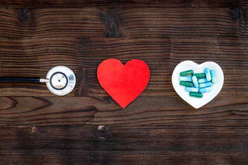 Treat heart. Pills in bowl in shape of heart near stethoscope on dark wooden background top view copyspace