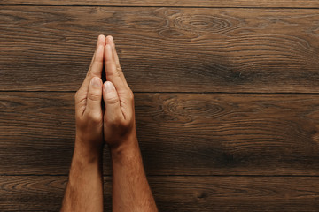 Male hands are folded in prayer on a wooden brown background. Copy space.