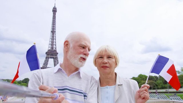 Senior couple with French flags near Eiffel Tower doing selfie on mobile with selfie-stick. Smiling tourist traveling in Europe. Active modern life after retirement. Family enjoying time together