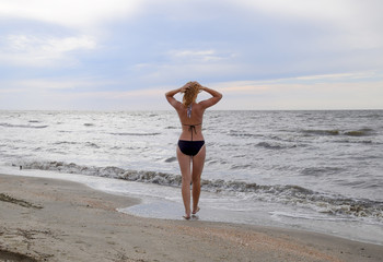Fototapeta na wymiar Girl in a bikini stands by the sea, a rear view. A beach walk of the blonde along the shore. The girl goes to the sea.