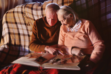 a married couple of retirement age sitting on a sofa by the fireplace wrapped in a plaid examines a family photo album.