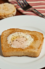 Single egg fried over light on white toast with the center  cut out, with fresh ground pepper, round, white dish, artisan fork and a red and white stripped towel in a basket 