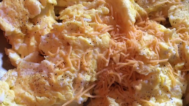 Cheddar cheese is sprinkled onto scrambled eggs from above in natural lighting.  
