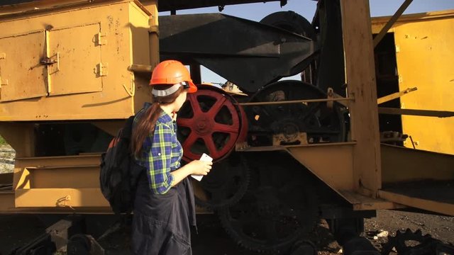 A young woman worker in orange helmet stands near a yellow old excavator and use a white telephone.