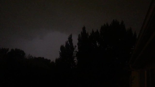 Lightning pulsates behind trees in summer storm before large flash encompasses sky at end.  