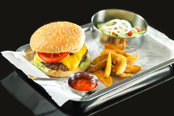Closeup fast food set including hamburger, ketchup, salad and french fries on a tray at black background.